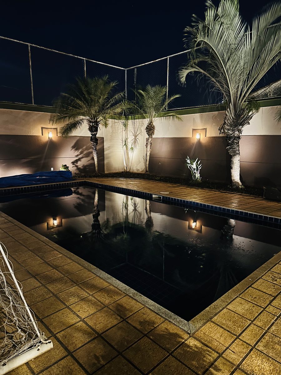 Área da piscina à noite, com palmeiras iluminadas por arandelas na parede e o reflexo delas na água.
