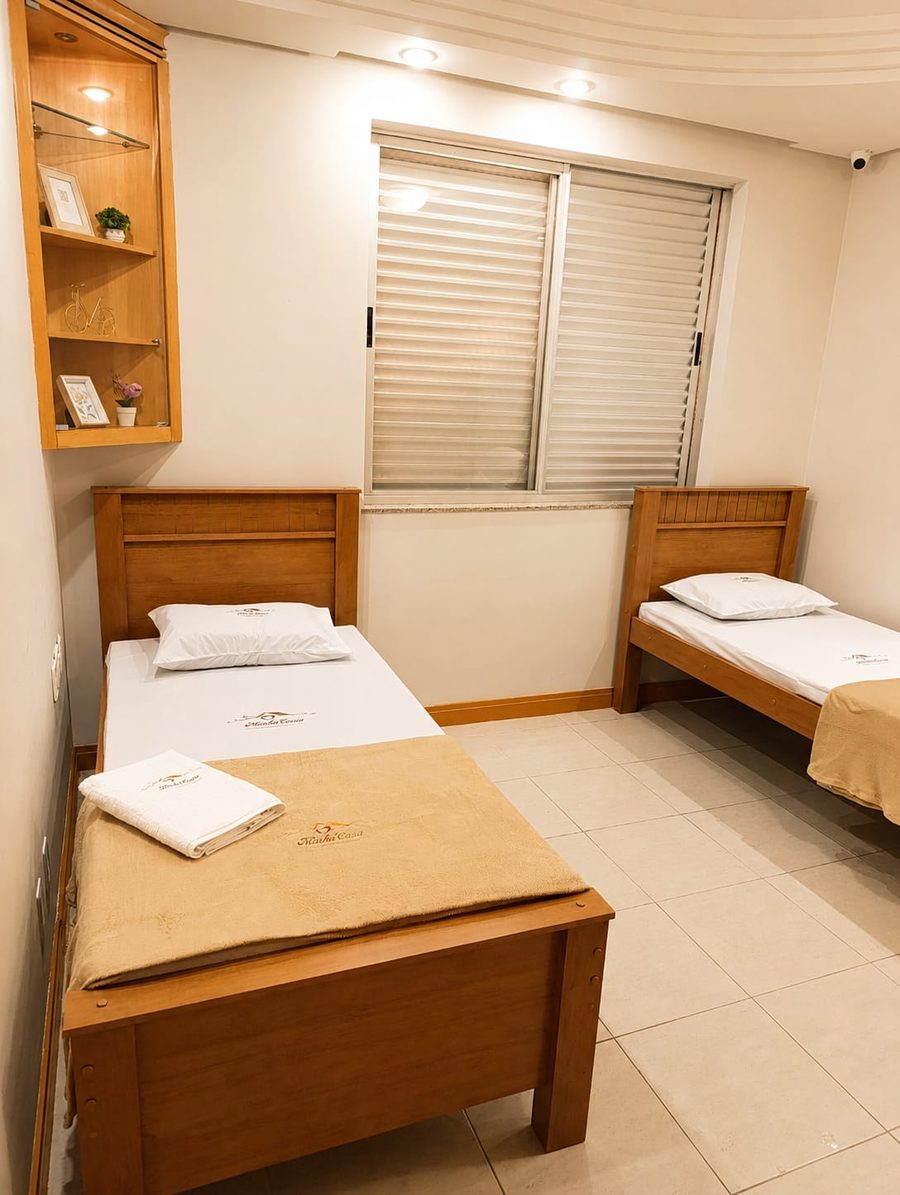 Quarto compartilhado com duas camas de madeira, roupa de cama bordada com a marca da casa e nicho de decoração iluminado.