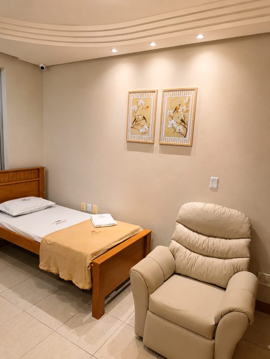 Quarto individual com cama de madeira, poltrona reclinável, quadros na parede e câmera de monitoramento no teto.